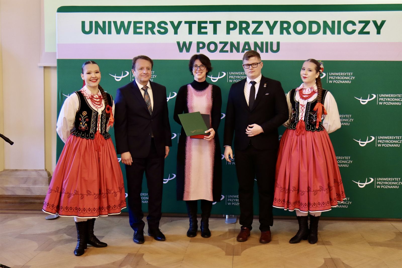5 osób, w środku dr Anna Szczepańska-Álvarez, po lewej jego magnificencja rektor  prof. dr hab. Krzysztof Szoskiewicz wraz z kobietą w stroju ludowym, po lewej  mężczyzna w garniturze i kobieta w stroju ludowym